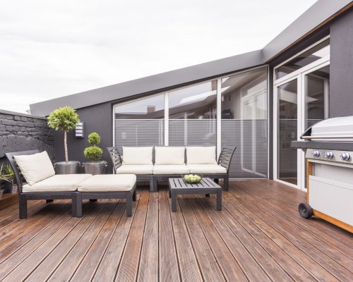 Bright garden furniture, grill and plants on cozy terrace with wooden floor and brick wall